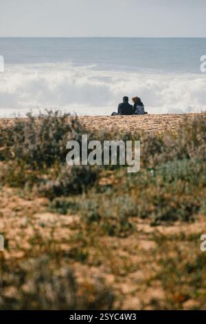 A couple sitting on the beach, watching the waves in Nazaré Stock Photo