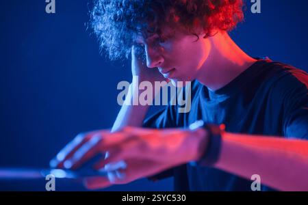 DJ is in the club. Young man with curly hair is indoors illuminated by neon lighting. Stock Photo