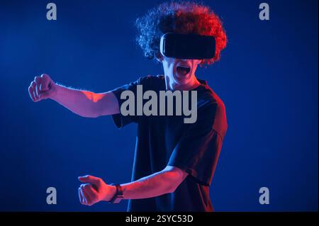 Racing game. In VR glasses. Young man with curly hair is indoors illuminated by neon lighting. Stock Photo