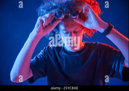 Taking off the retro style glasses. Young man with curly hair is indoors illuminated by neon lighting. Stock Photo
