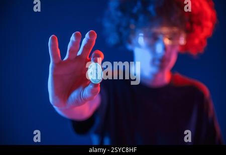 Holding bitcoin. Young man with curly hair is indoors illuminated by neon lighting. Stock Photo
