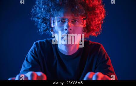 Gamer is focused by playing. Young man with curly hair is indoors illuminated by neon lighting. Stock Photo