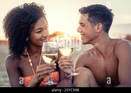 Young interracial couple enjoying a romantic sunset on the beach, toasting with glasses of white wine, sharing smiles and a joyful summer moment. Stock Photo