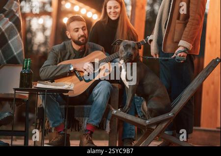 Group of people is having fun together. Man playing guitar, his friend is playing with dog. Stock Photo