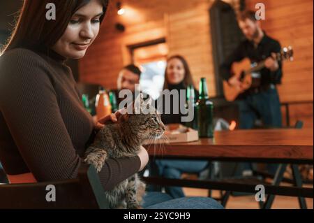 Woman with cat on her legs. Group of friends is having good weekend indoors in the wooden building together. Stock Photo