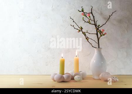 Easter decoration in pastel rose and yellow colors, quince branch in a vase, two candles and speckled eggs on a wooden sideboard against a light rusti Stock Photo