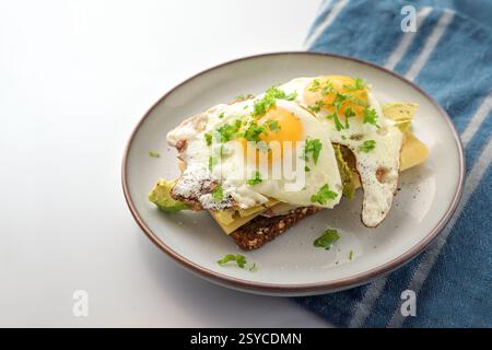Fried eggs on dark rye bread with cheese, avocado and parsley garnish, hearty meal for breakfast, lunch or dinner, blue kitchen towel, light gray back Stock Photo
