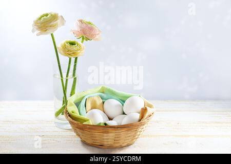 Easter decoration in pastel colors, white eggs in a small basket with a napkin and a glass vase with ranunculus flowers on a light wooden table, copy Stock Photo