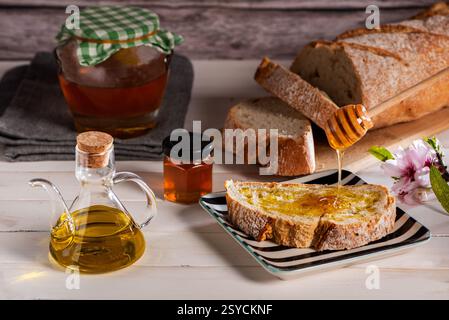 Toasted slice of bread with olive oil and honey, on a white table with an oil dispenser, a jar of honey, and a loaf of bread sliced. Stock Photo