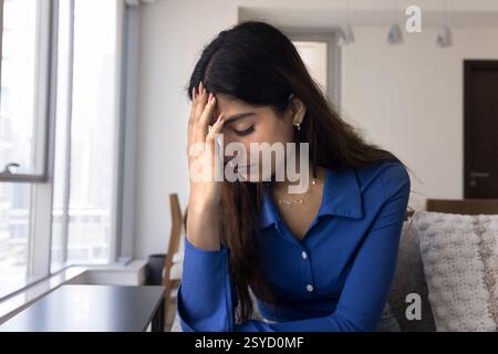Young Indian woman sit on sofa looks stressed and worried Stock Photo