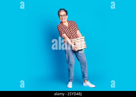 Stylish mature woman holding stack of books, smiling confidently against a vibrant blue background, posing in colorful casual outfit Stock Photo