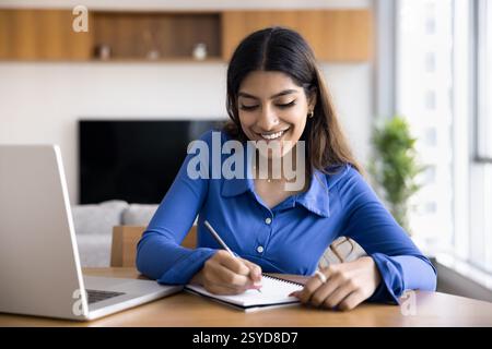 Woman smiles, writes in notebook seated at desk with laptop Stock Photo