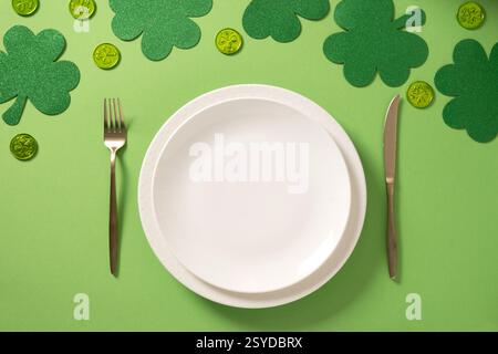 St Patrick's Day table setting with empty white plate and traditional shamrock, clover leaves and coins on light green background. View from above. Stock Photo