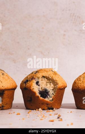 Three blueberry muffins in a row with the center one showing a bite taken out, revealing its moist interior texture with visible berries. Stock Photo