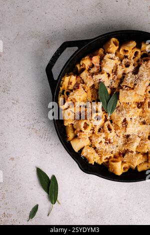 A rustic cast iron skillet filled with creamy rigatoni pasta, crispy pancetta, and parmesan, garnished with fresh sage on a textured neutral backgroun Stock Photo