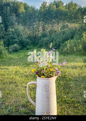 A white textured ceramic jug filled with fresh wildflowers sits on green grass, with a dense forest in the background. Photographed in natural dayligh Stock Photo