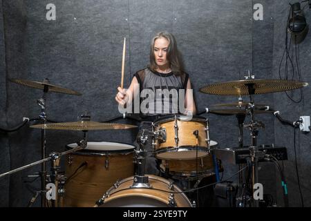 Female drummer playing drum kit in soundproof studio. Focused musician holding drumsticks, performing energetic beat. Professional music rehearsal and practice session. Stock Photo