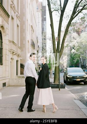 Pair strolls hand in hand down a city street, dressed elegantly for a special occasion during springtime. Stock Photo