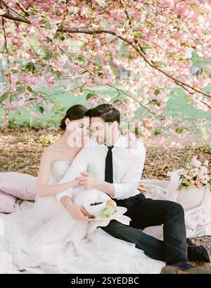 A newlywed couple shares a sweet moment while sitting on a blanket surrounded by blooming cherry blossoms. Stock Photo