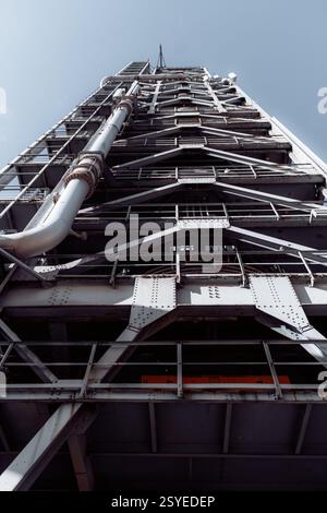 Low-angle view of an industrial metal structure with steel beams, rivets, railings, and large pipes against a clear sky. Rust and weathering visible o Stock Photo
