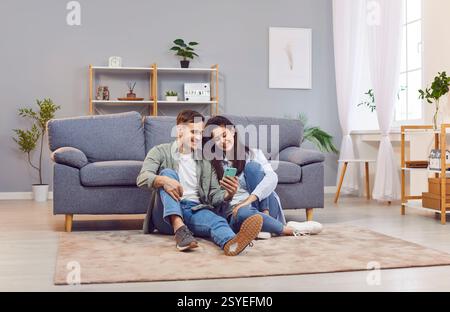 Happy couple at home, young woman and man floor sitting close in new living room, using smartphone Stock Photo