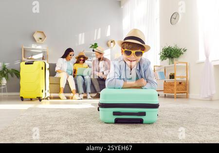 Boy with suitcase looking at camera while his family booking hotel online using laptop. Stock Photo