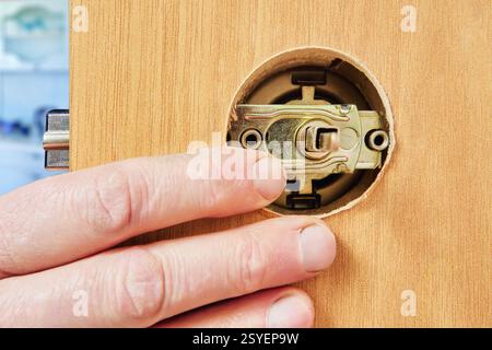 Brass latch hardware is positioned inside pre-cut hole in wooden door, with hand steadying door edge during assembly. Stock Photo