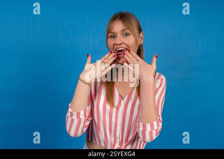 Young woman with long blond hair wearing pink and white striped shirt expressing surprise covering her mouth with hands Stock Photo