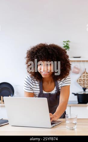Happy young woman in apron using laptop while cooking in the kitchen Stock Photo