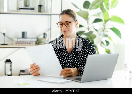 Focused confident businesswoman, company manager, sitting at the desk with a laptop, holding financial report, concentrated looking at the graphs and analyzing data in a modern office Stock Photo