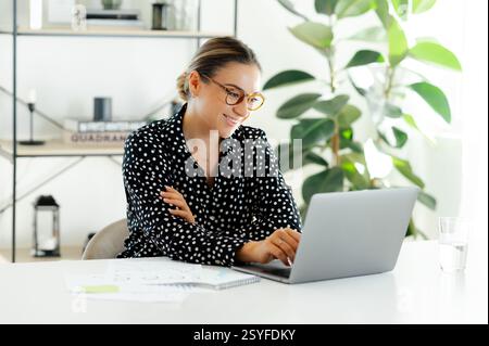 Focused confident caucasian businesswoman in glasses, sitting at the work desk with laptop, typing on keyboard, smiling and working on a financial report in a modern office Stock Photo
