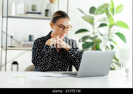 Confident positive businesswoman with glasses, sitting at the desk with laptop, reading information on a laptop screen, smiling and working with financial data in a bright modern office Stock Photo