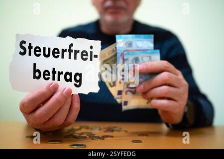 Munich, Bavaria, Germany - February 27, 2025: PHOTOMONTAGE. An elderly man is holding a piece of paper with the words Steuerfreibetrag and several euro banknotes. There are coins on the table. The picture focuses on financial aspects such as taxes, tax allowances and pension income. *** FOTOMONTAGE. Ein älterer Mann hält einen Zettel mit der Aufschrift Steuerfreibetrag sowie mehrere Euro-Geldscheine in der Hand. Auf dem Tisch liegen Münzen. Das Bild thematisiert finanzielle Aspekte wie Steuern, Freibeträge und Renteneinkommen. Stock Photo