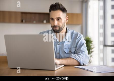 Handsome focused man sits at desk using laptop Stock Photo