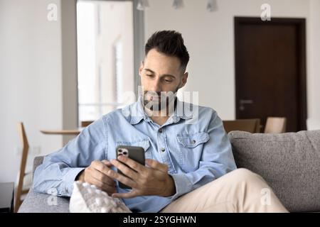 Hispanic man using his smartphone seated on sofa at home Stock Photo