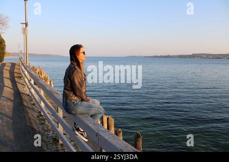 Eine junge Frau sitzt entspannt auf einem Geländer an der Uferpromenade und blickt in die Ferne über das ruhige, tiefblaue Wasser des Bodensees. Stock Photo