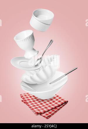 Clean dishware and silverware flying over red checkered napkin on pink background Stock Photo