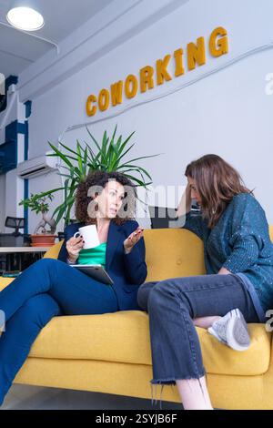 Two businesswomen are having a serious conversation on a yellow sofa in a coworking space Stock Photo