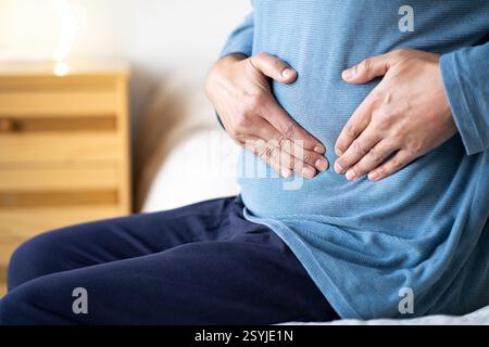 Man sitting alone on his bed and suffering from stomach cramps while home alone.Abdominal pain can be caused by digestive problems, infections, intest Stock Photo