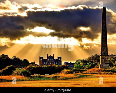 Seaton Delaval Hall and obelisk in corn fields at sunset with dark  clouds and sunbeams bursting through Stock Photo