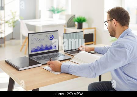 Focused trader sits at desk looking at laptop screen monitors for stock market data Stock Photo