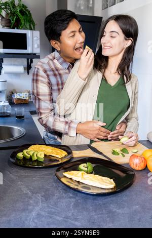 Happy couple sharing a playful moment while preparing breakfast in the kitchen. the man hugs the woman from behind and feeds her an avocado slice. pla Stock Photo