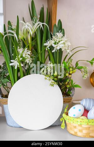 Decorative pot filled with lush green plants, white flowers and colorful Easter eggs in basket on a shelf with empty circle, blank round display cardb Stock Photo