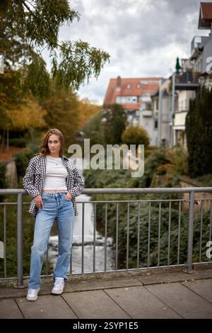 Autumn Urban Serenity: Beautiful Young Woman in Checkered Shirt and Crop Top Poses on Bridge with Vibrant Foliage and Red-Roofed Buildings. Casual Fas Stock Photo