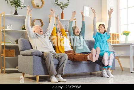 Happy elderly people with nurse sitting on couch, raising hands, cheering and enjoying activity. Stock Photo