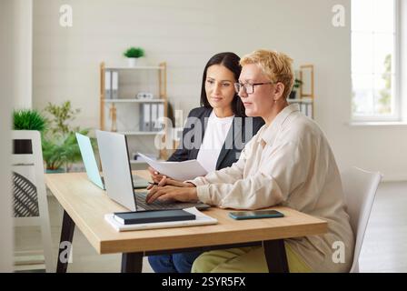 Two female colleagues working together at desk in office, using laptop and analyzing business data. Stock Photo