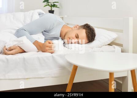 Young man sleeping well in bed on soft white pillow holding mug, resting, good night sleep Stock Photo
