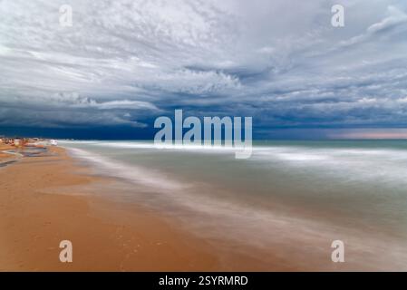 A long exposure captures the motion of the waves as they meet the shore under a dramatic, cloudy sky at the beach. Cut-off low Stock Photo