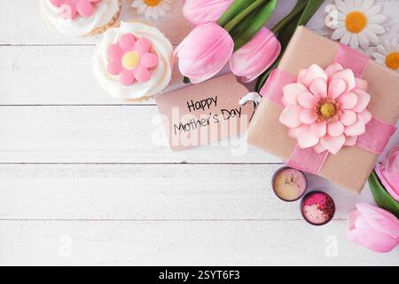 Happy Mothers Day corner border with gift tag and box, flowers, cupcakes and chocolate. Overhead view against a white wood background. Copy space. Stock Photo