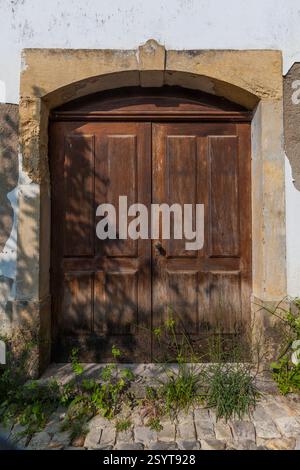 A weathered double wooden door with a stone archway is set in a white wall, with weeds growing at its base Stock Photo